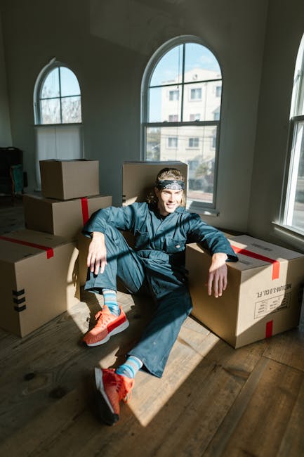 A person, dressed in casual clothing with a white t-shirt, jeans, and a black cap, is seen carrying three cardboard boxes during a home relocation process in front of a white delivery van. The van, pa